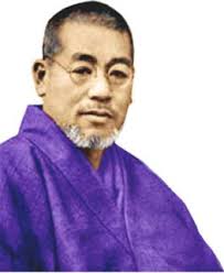 An elderly man in glasses and purple robe, gazing forward with a calm expression, against a plain background.