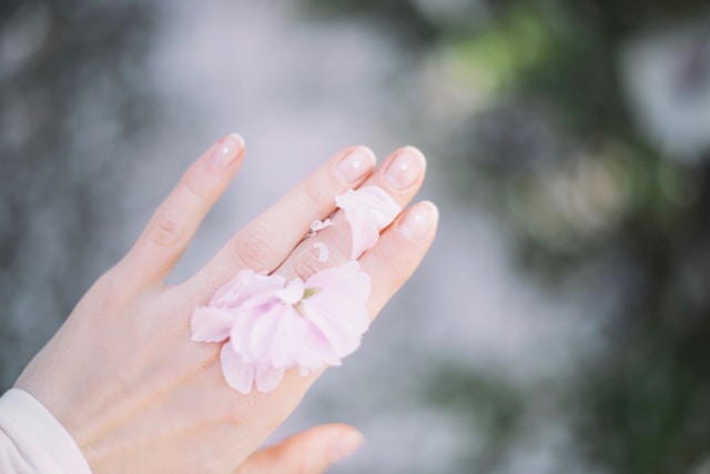 Hand with delicate pale pink flower petals on fingers, set against a soft-focus green and white background.